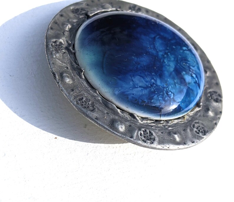 May include: A round pewter brooch with a blue and white glass cabochon in the center. The pewter has a textured surface and is decorated with raised designs.