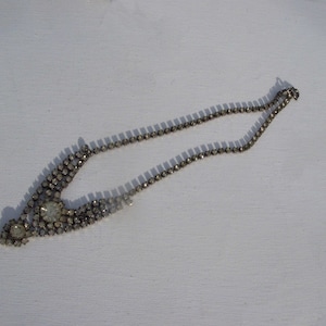 Pode incluir: Um colar prateado com strass redondos e transparentes. O colar apresenta um design central com pedras maiores e uma corrente delicada. Está sobre um fundo branco, realçando o seu brilho e design.