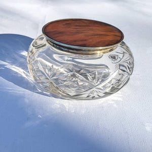 May include: A clear, cut-glass jar with a wooden lid. The jar has a silver rim and a starburst pattern on the sides. The lid is oval-shaped and has a dark brown wood grain.