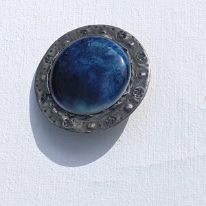May include: A round, silver-toned brooch with a blue and white glass cabochon in the center. The cabochon has a swirling pattern.