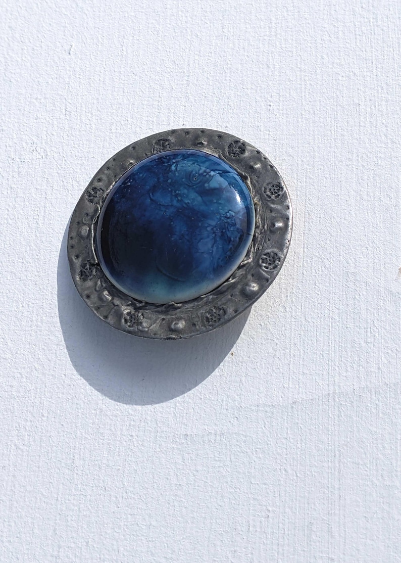 May include: A round, silver-toned brooch with a blue and white glass cabochon in the center. The cabochon has a swirling pattern.