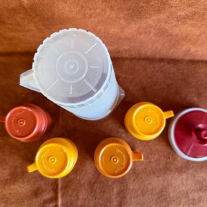 Könnte beinhalten: Ein Set aus Kunststoff-Küchenutensilien, bestehend aus einem großen, durchscheinenden Krug mit Deckel und fünf kleineren Behältern mit Deckeln in Orange-, Gelb- und Dunkelrottönen. Jeder Deckel ist mit einem kleinen Griff versehen.