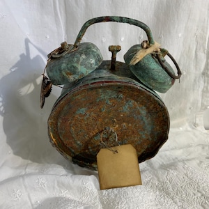 May include: A vintage alarm clock with a distressed, rusty appearance. The clock has a green and brown patina, with a curved handle and two bells on top. A blank tag hangs from the front. The clock is a unique decorative item.