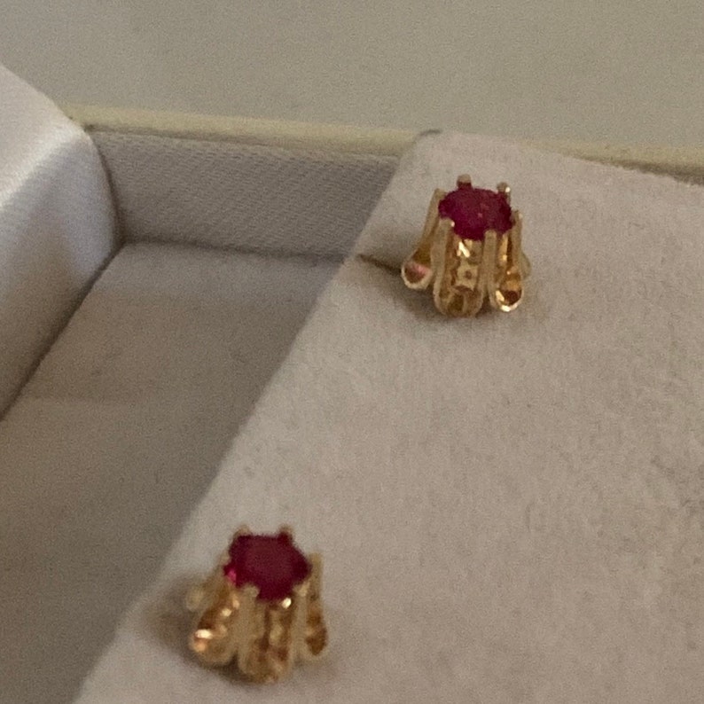 May include: A pair of gold earrings with red gemstone accents. The earrings have a detailed design and are displayed in a white box.