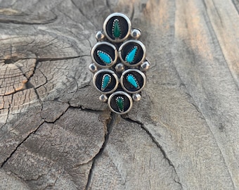 May include: A silver-toned ring with an oval design. The ring features six circular settings, each containing a gemstone. The gemstones alternate between turquoise and green, creating a striking contrast. The ring is set against a rustic wooden surface.