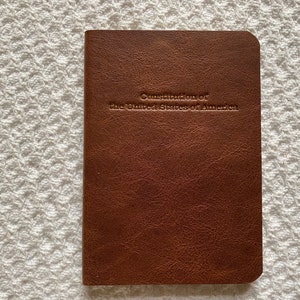 May include: A brown leather-bound book with rounded corners. The embossed text on the cover reads "Constitution of the United States of America". The book is closed and lies on a white, textured surface.