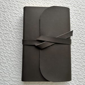 May include: A black leather journal with a flap closure and a leather strap that wraps around the front and ties in a knot.