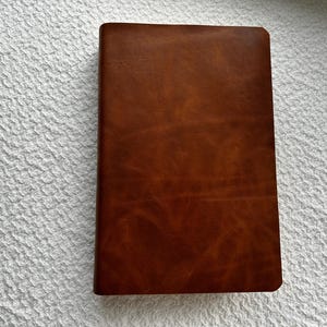 May include: A rectangular notebook with a smooth, medium-brown leather cover featuring subtle natural grain variations and a consistent, matte finish. The front cover is plain and unadorned, showcasing the rich, warm tones of the material.