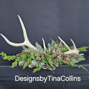 May include: A rustic centerpiece featuring deer antlers, evergreen branches, pinecones, and feathers. The arrangement is set against a dark background and includes the text "DesignsbyTinaCollins".