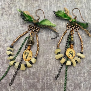 May include: A pair of handmade earrings with a steampunk aesthetic. Each earring features a copper-colored hook, a decorative top, and dangling elements including cream-colored stones, black beads, and a chain. One earring has the number "7 IN" and the other has the number "8".