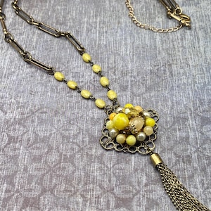 May include: Vintage gold-tone necklace featuring a decorative pendant with yellow beads and a tassel. The necklace has a chain with large rectangular links and a smaller chain for adjusting the length. The pendant has a filigree design.