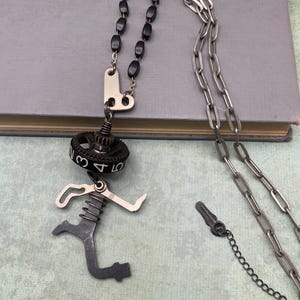 May include: A necklace featuring a silver-toned chain, black beads, and a pendant assembly. The pendant includes a dark gear with the numbers 3, 4, and 5 visible, connected to a metallic, spring-loaded, angular component resembling a mechanical part.