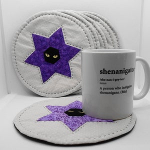 May include: Set of six purple and white star-shaped fabric coasters with a black cat face in the center. The coasters are on a white surface with a white mug that says "shenanigator /she-nan-i-gey-ter/ noun A person who instigates shenanigans. (Me)"