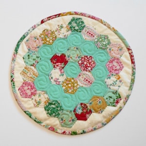 May include: A round, quilted coaster with a hexagonal pattern in shades of pink, red, green, and yellow. The center of the coaster is a turquoise hexagon with a swirl design.
