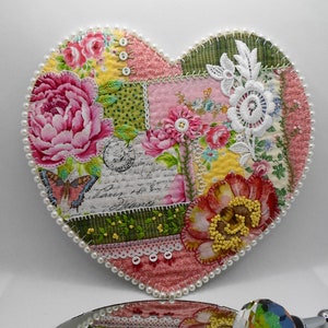 May include: Heart-shaped decorative piece with a patchwork design. It features floral patterns, a butterfly, and script details. The edges are trimmed with small white pearls. The piece is embellished with lace and buttons.