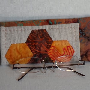 May include: A handmade fabric glasses case with a geometric pattern of orange, brown, and red hexagons. The case is made of a light brown fabric with a white lining. A pair of clear glasses is resting on top of the case.
