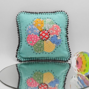 May include: A square, light blue pincushion with a floral design made of colorful fabric patches. The patches are arranged in a flower shape and are stitched onto the cushion. The edges are trimmed with black beads.