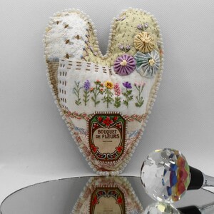 May include: A heart-shaped fabric ornament with embroidered flowers and a label that reads "Bouquet de Fleurs". The ornament is decorated with white beads around the edges.