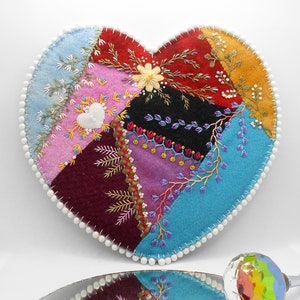 May include: A heart-shaped patchwork quilt with a variety of colors and textures. The quilt is made of felt and is decorated with embroidery. The quilt is surrounded by a white beaded trim.