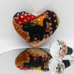 May include: A heart-shaped felt ornament with a black cat silhouette embroidered on a patchwork of colorful fabrics. The ornament is hanging from a white ribbon and is reflected in a mirror.