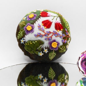 May include: A felt ball ornament with a whimsical embroidered design featuring a frog, flowers, and leaves in shades of green, red, yellow, purple, and white.