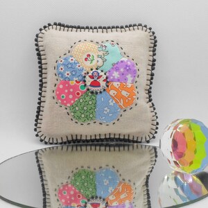 May include: A beige fabric pillow with a black beaded border features a colorful patchwork flower design. The flower is made of various fabrics in shades of pink, blue, green, yellow, and orange.