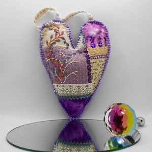 May include: Purple and cream heart-shaped fabric ornament with embroidered flowers and a handwritten message. The ornament is hanging from a loop of fabric and is reflected in a mirror.