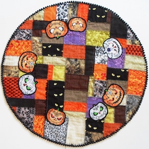 May include: A round, patchwork Halloween tree skirt with a black beaded edge. The skirt features various fabric squares in orange, brown, purple, and black. Appliqued sugar skull and jack-o'-lantern designs are scattered across the surface, with cat eyes peeking out from the black squares.