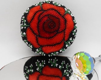 May include: A red felt rose with green leaves and white beads is displayed on a black background. The rose is reflected in a mirror.