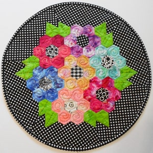 May include: A round table runner with a black background and white polka dots. A colorful flower design is in the center of the runner, made up of six hexagonal flowers in shades of pink, red, blue, purple, orange, and green.