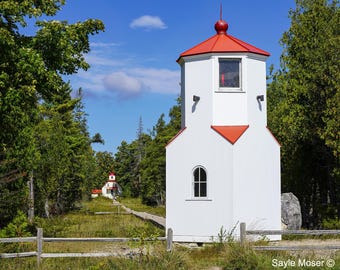 May include: A white lighthouse with a red roof and a red trim. The lighthouse is surrounded by trees and a wooden fence. The lighthouse is located on a grassy area.