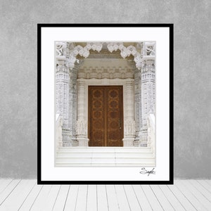 May include: A white marble temple entrance with a large, intricately carved wooden door. The door is made of two panels and has a symmetrical design with floral motifs. The temple is decorated with intricate carvings and details.
