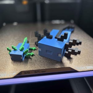 May include: Two blocky, pixelated axolotl figures. The smaller one is blue with bright green gills and fins. The larger one is blue with dark brown gills and fins. Both figures feature a square head, rectangular body, and small eyes.