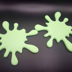May include: Two light green coasters shaped like liquid paint splatters. Each coaster features a central circular area with multiple irregular, rounded protrusions extending outward in a radial pattern, mimicking the appearance of a splash.