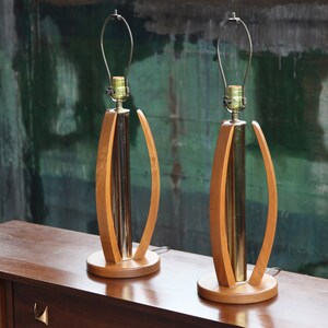 Pode incluir: Um par de candeeiros de mesa de estilo moderno de meados do s&eacute;culo com bases de madeira e detalhes em lat&atilde;o. Os candeeiros t&ecirc;m um design curvo &uacute;nico e est&atilde;o em excelente estado.