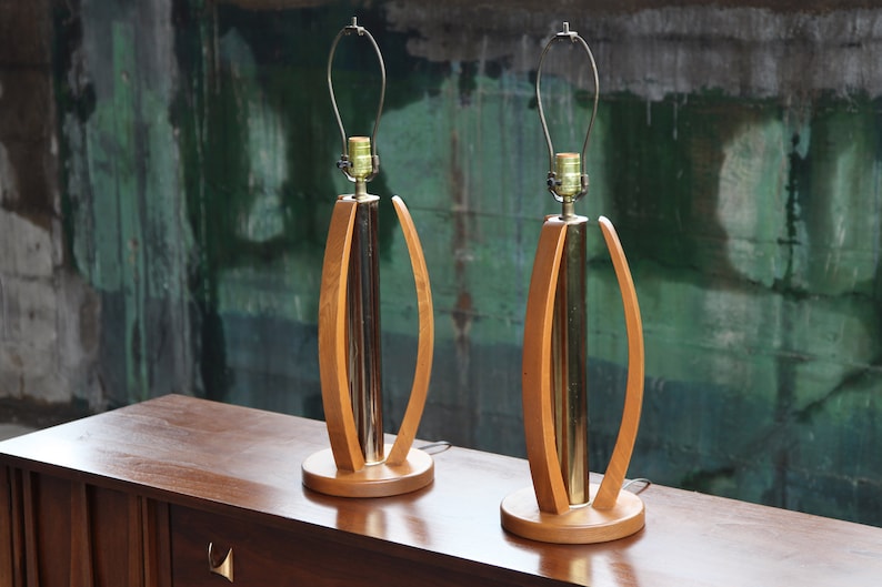 Pode incluir: Um par de candeeiros de mesa de estilo moderno de meados do s&eacute;culo com bases de madeira e detalhes em lat&atilde;o. Os candeeiros t&ecirc;m um design curvo &uacute;nico e est&atilde;o em excelente estado.