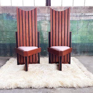 May include: Two tall, wooden chairs with black accents and a light pink cushion. The chairs are on a white fluffy rug in front of a green and gray wall.