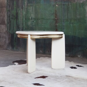 May include: White side table with a gold trim around the top edge. The table has a rounded, oval-shaped top and four legs.