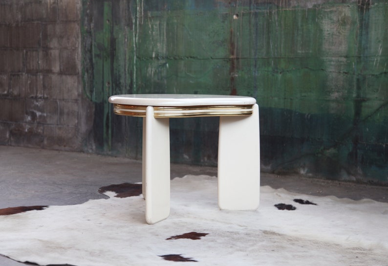May include: White side table with a gold trim around the top edge. The table has a rounded, oval-shaped top and four legs.