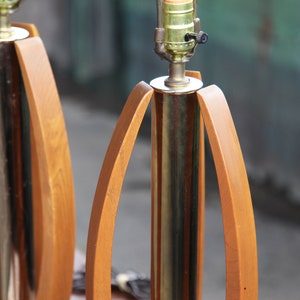 Pode incluir: Um par de candeeiros de mesa de estilo moderno de meados do s&eacute;culo com base em lat&atilde;o e detalhes em madeira. Os candeeiros t&ecirc;m um design &uacute;nico com bra&ccedil;os de madeira curvos que criam uma forma geom&eacute;trica.