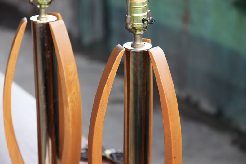 Pode incluir: Um par de candeeiros de mesa de estilo moderno de meados do s&eacute;culo com base em lat&atilde;o e detalhes em madeira. Os candeeiros t&ecirc;m um design &uacute;nico com bra&ccedil;os de madeira curvos que criam uma forma geom&eacute;trica.