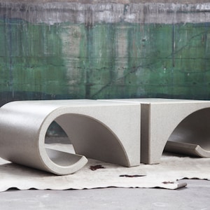 May include: Two gray concrete coffee tables with a unique curved design. The tables are placed on a white fur rug in front of a green wall.