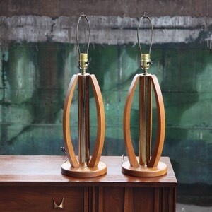Pode incluir: Um par de candeeiros de mesa de estilo moderno de meados do s&eacute;culo, em madeira e lat&atilde;o. Os candeeiros t&ecirc;m um design oval &uacute;nico com um cilindro de lat&atilde;o no centro.