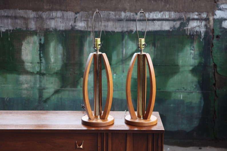 Pode incluir: Um par de candeeiros de mesa de estilo moderno de meados do s&eacute;culo, em madeira e lat&atilde;o. Os candeeiros t&ecirc;m um design oval &uacute;nico com um cilindro de lat&atilde;o no centro.