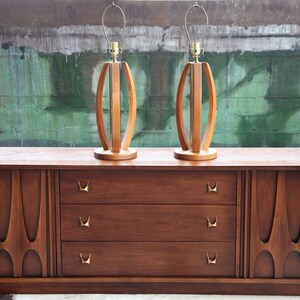 Pode incluir: Uma c&oacute;moda de estilo moderno de meados do s&eacute;culo com tr&ecirc;s gavetas e dois arm&aacute;rios laterais. A c&oacute;moda &eacute; feita de madeira e tem um acabamento castanho escuro. Duas l&acirc;mpadas de madeira com um design curvo &uacute;nico s&atilde;o colocadas no topo da c&oacute;moda.