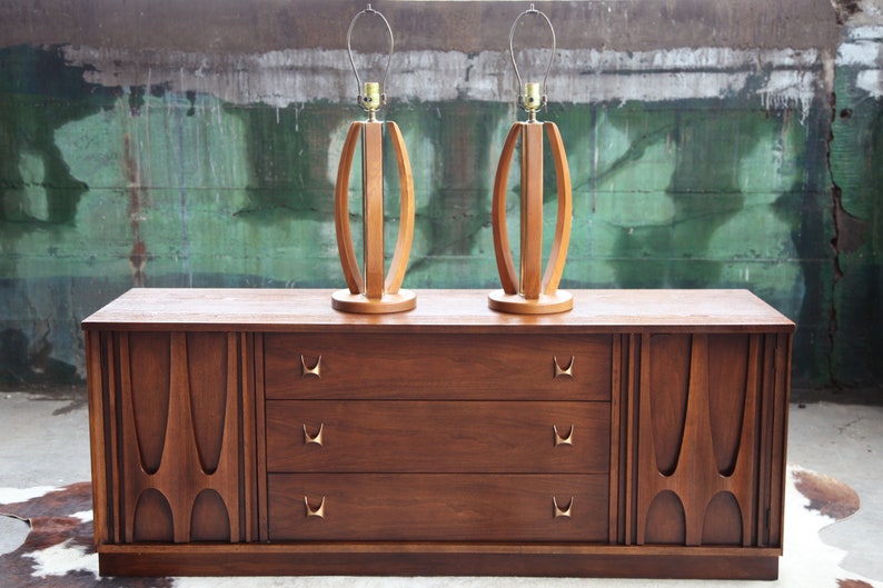 Pode incluir: Uma c&oacute;moda de estilo moderno de meados do s&eacute;culo com tr&ecirc;s gavetas e dois arm&aacute;rios laterais. A c&oacute;moda &eacute; feita de madeira e tem um acabamento castanho escuro. Duas l&acirc;mpadas de madeira com um design curvo &uacute;nico s&atilde;o colocadas no topo da c&oacute;moda.