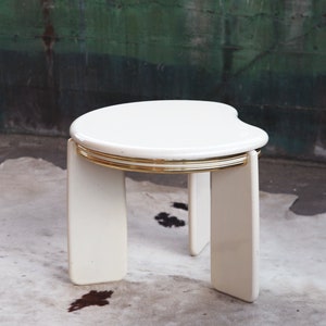 May include: White side table with a rounded, asymmetrical top and gold trim. The table has four legs and is made of a smooth, white material.