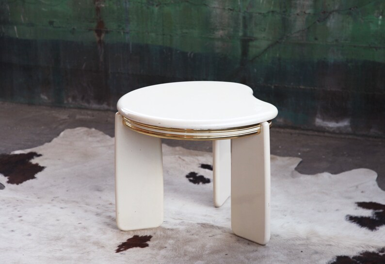 May include: White side table with a rounded, asymmetrical top and gold trim. The table has four legs and is made of a smooth, white material.