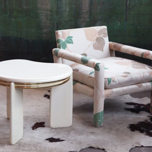 May include: A white side table with a gold trim and a floral patterned armchair. The table has a unique, curved top and three legs. The chair has a light brown and green floral pattern on the fabric.