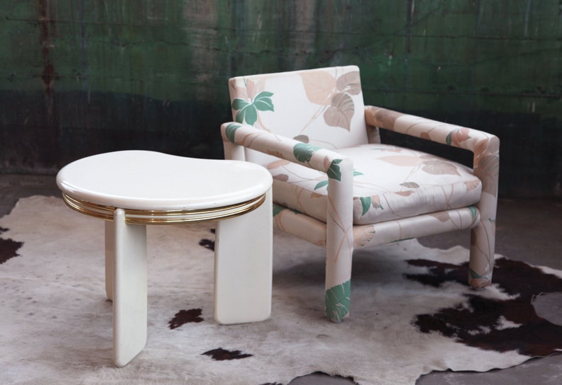 May include: A white side table with a gold trim and a floral patterned armchair. The table has a unique, curved top and three legs. The chair has a light brown and green floral pattern on the fabric.